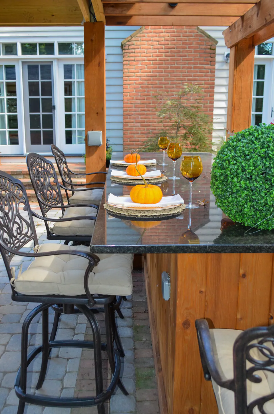 Close view of outdoor bar with dining chairs, wood counter, pergola post, paver patio, and house doors.